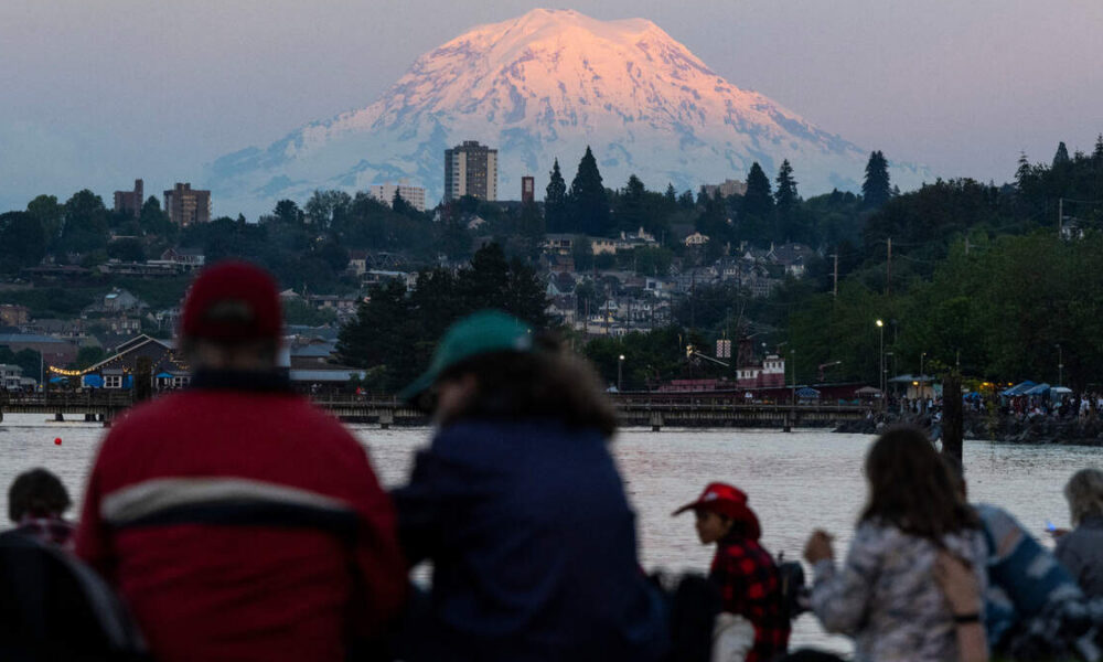 People gathered by a waterfront watching the scene with a pink-glow snow-capped mountain in the background at sunset/dusk.