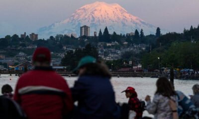 People gathered by a waterfront watching the scene with a pink-glow snow-capped mountain in the background at sunset/dusk.