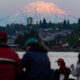 People gathered by a waterfront watching the scene with a pink-glow snow-capped mountain in the background at sunset/dusk.