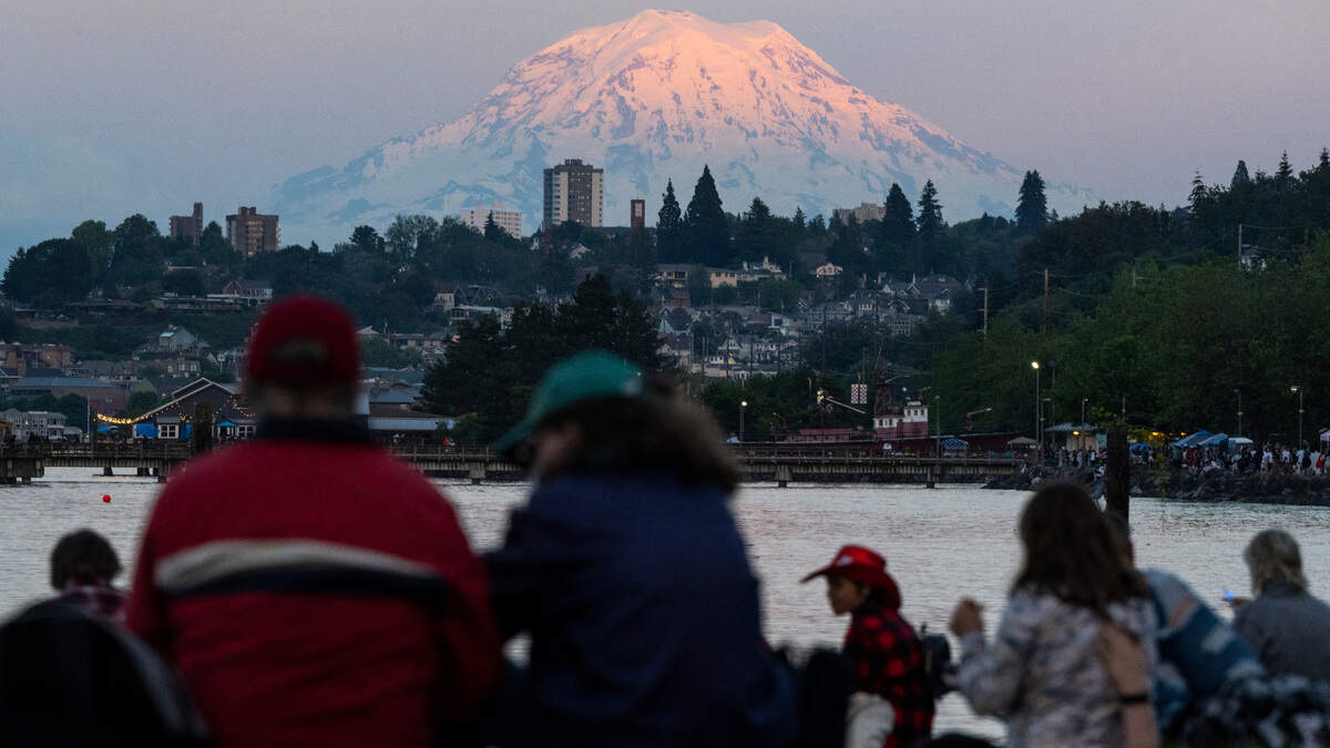 People gathered by a waterfront watching the scene with a pink-glow snow-capped mountain in the background at sunset/dusk.