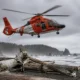 US Coast Guard helicopter in bright orange hovering above a driftwood-strewn rocky beach as gray waves crash and a forested cliffline looms in the background.
