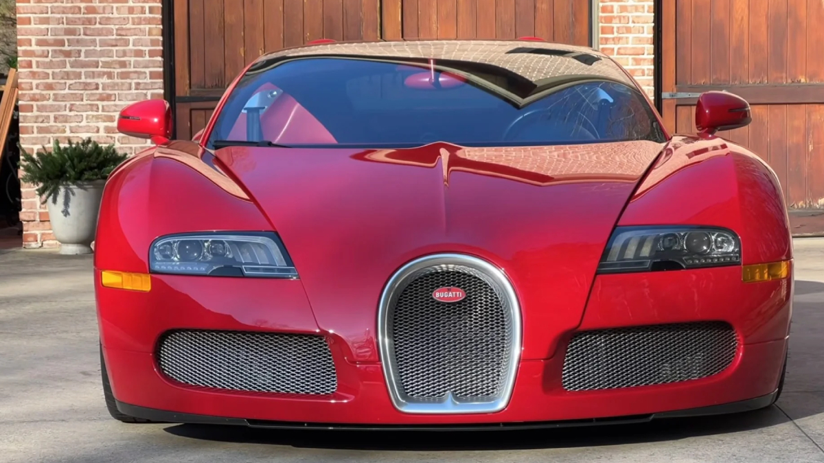 Front view of a red Bugatti sports car parked on a driveway with brick walls in the background, showing a distinctive horseshoe grille and sleek headlamps.