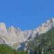 Rocky mountain peaks rise above a dense green pine forest under a bright blue sky in an alpine landscape.