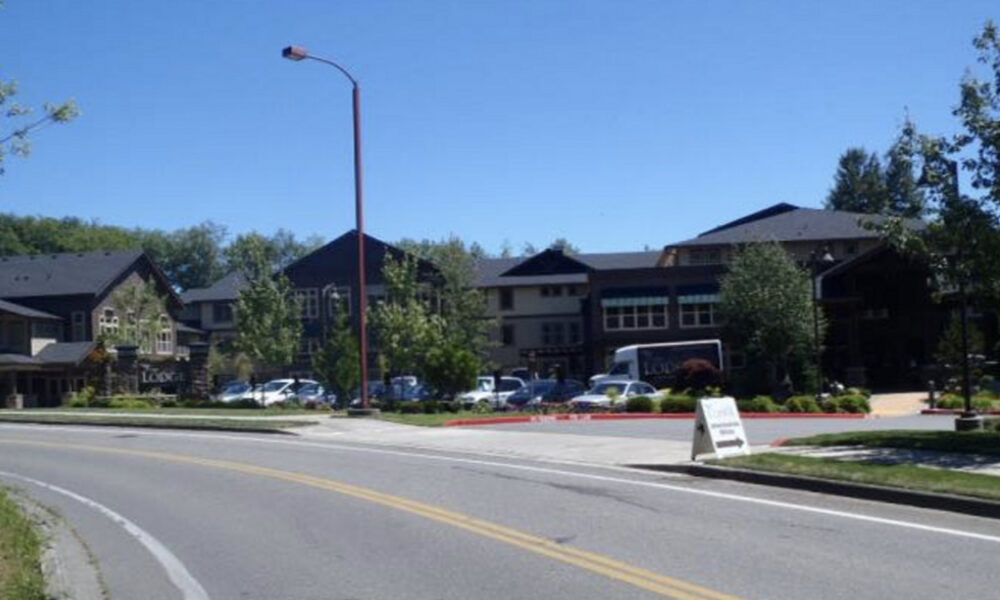Row of modern storefronts and a parking lot along a curved road under a clear blue sky, with several cars parked outside the buildings.