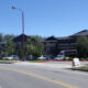 Row of modern storefronts and a parking lot along a curved road under a clear blue sky, with several cars parked outside the buildings.