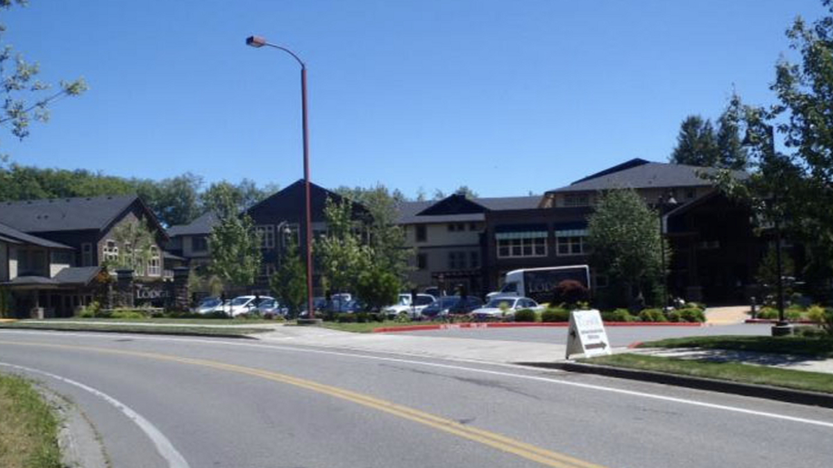 Row of modern storefronts and a parking lot along a curved road under a clear blue sky, with several cars parked outside the buildings.