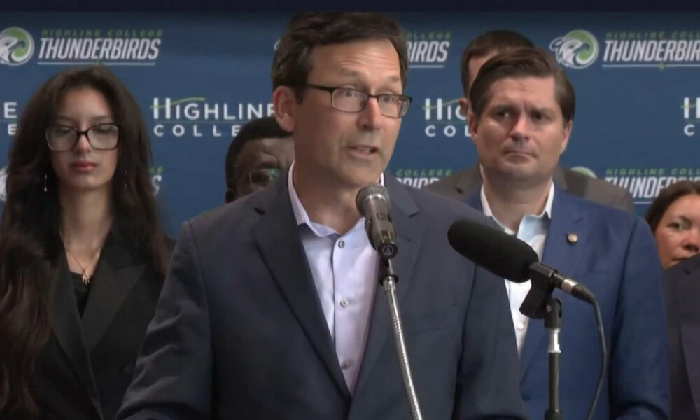 Man in a navy suit speaks at a podium with two microphones during a press conference; backdrop shows Highline College and Thunderbirds logos.