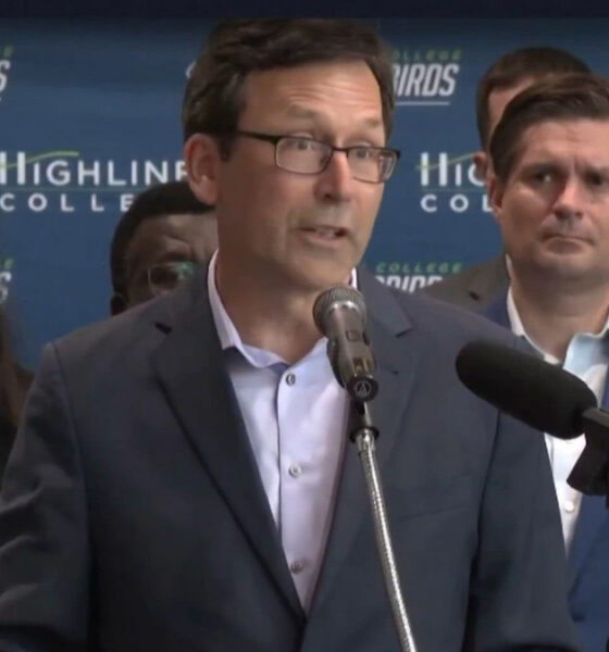 Man in a navy suit speaks at a podium with two microphones during a press conference; backdrop shows Highline College and Thunderbirds logos.