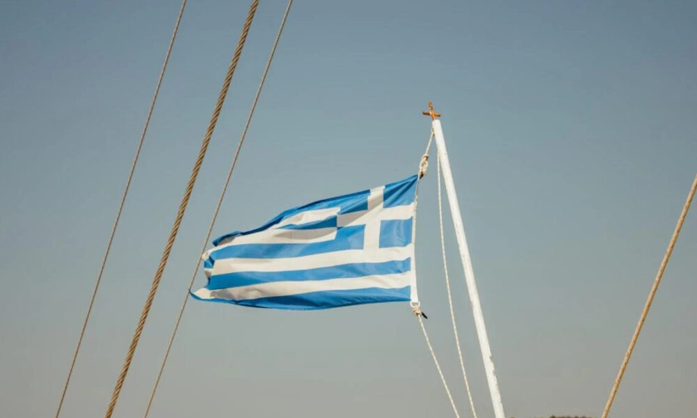 Blue and white Greek flag flying from a sailboat mast against a clear blue sky, with rigging cables nearby.