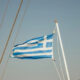 Greek flag fluttering on a sailboat mast against a clear blue sky, with rigging ropes nearby.