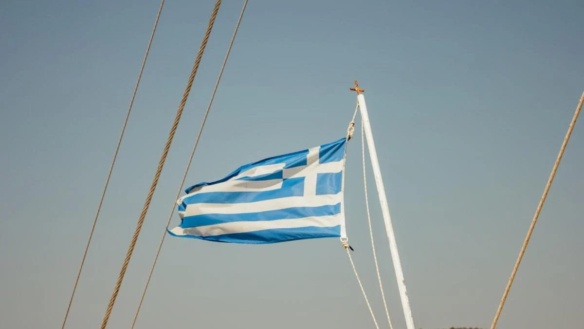 Greek flag fluttering on a sailboat mast against a clear blue sky, with rigging ropes nearby.