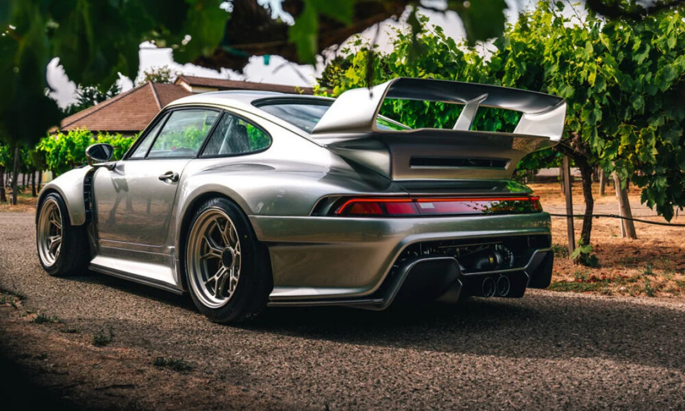Rear three-quarter view of a silver sports car with a large rear wing parked on a gravel path among vineyard rows.