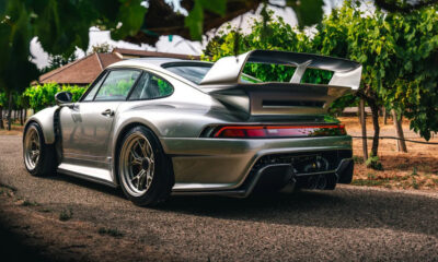 Rear three-quarter view of a silver sports car with a large rear wing parked on a gravel path among vineyard rows.