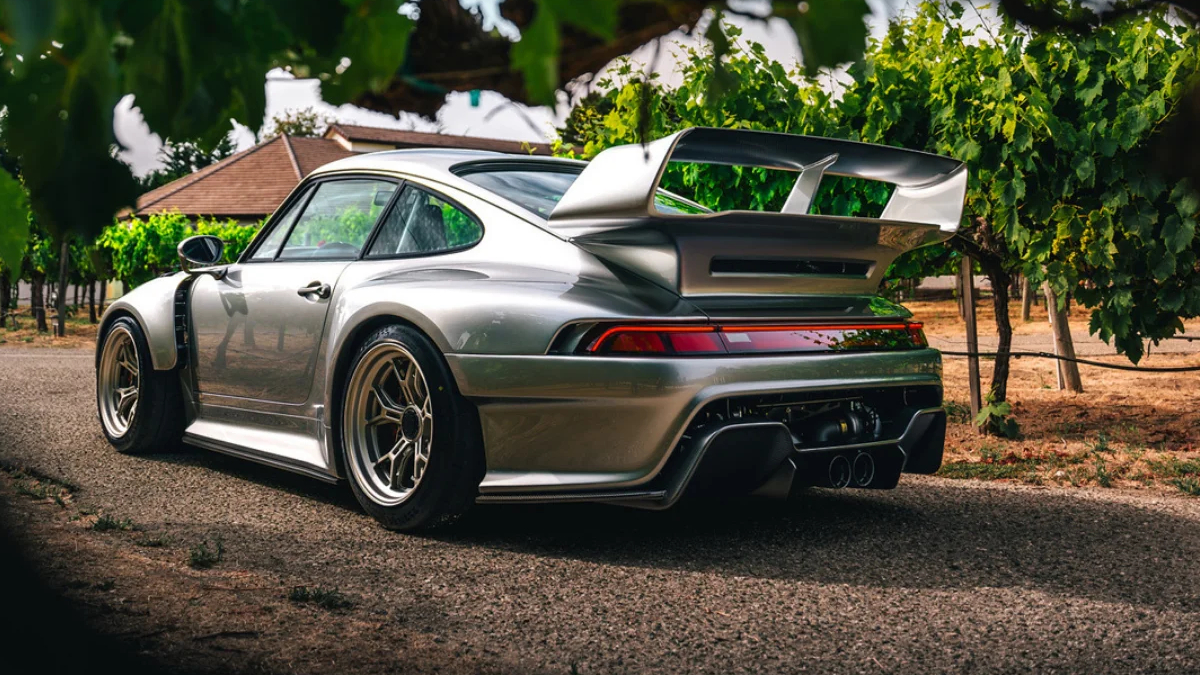 Rear three-quarter view of a silver sports car with a large rear wing parked on a gravel path among vineyard rows.
