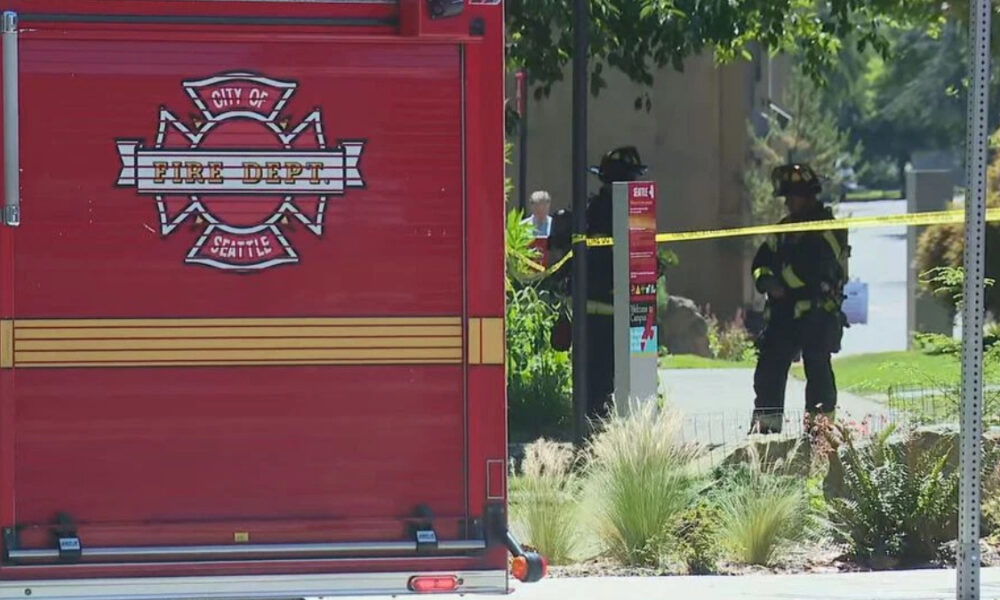 Rear view of a red City of Seattle Fire Department engine with two firefighters behind yellow caution tape in a suburban street.