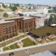 A university campus plaza with a red brick building and a large brown-roofed cafe, trees, and people walking nearby.