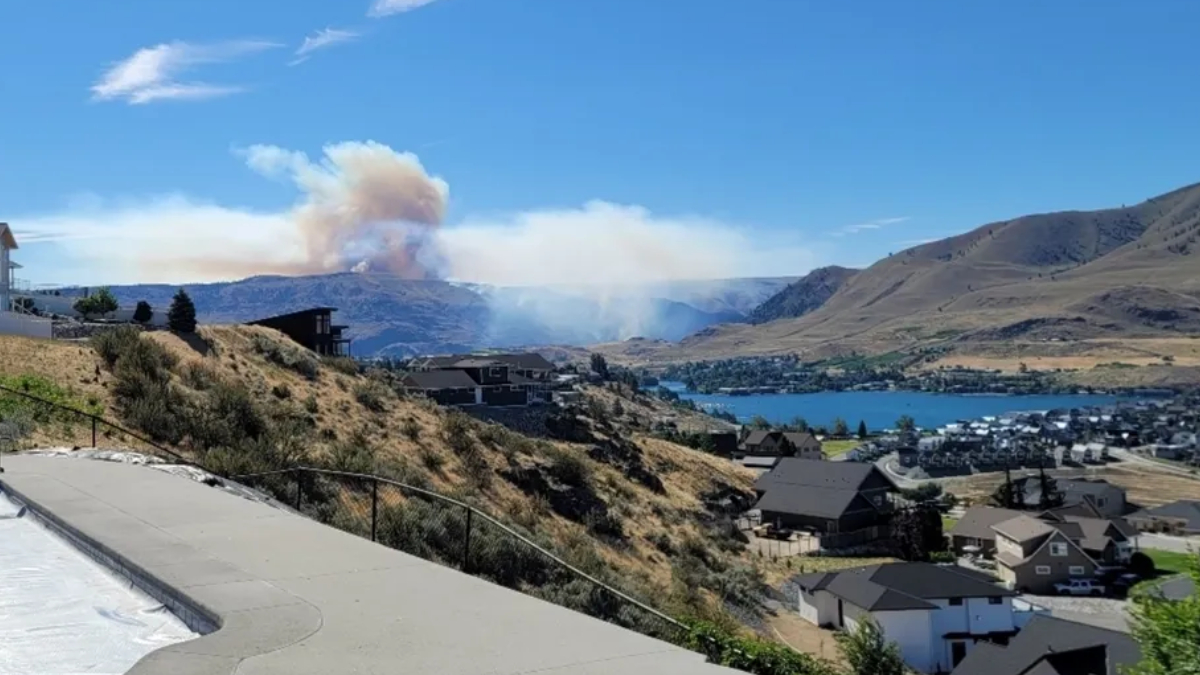 Hillside residential area overlooking a blue lake with a large smoke plume rising from a distant wildfire in the mountains under a clear sky, 2.