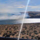 Desert-like shoreline overlooking a bright lake with rolling hills and a diagonal sun flare across the scene.