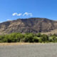 Wide landscape of arid mountains under a bright blue sky, with a line of green shrubs in the foreground.