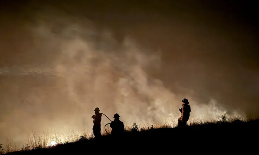 Silhouetted firefighters on a hill at night, battling a fire with thick smoke behind them.