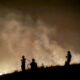Silhouetted firefighters on a hill at night, battling a fire with thick smoke behind them.