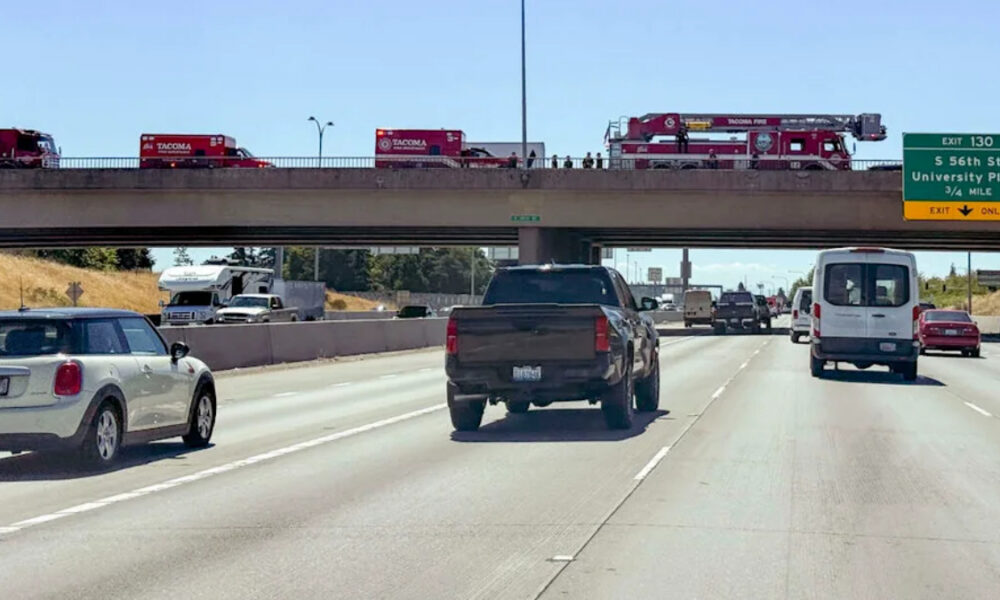Fire trucks lined up on an overpass above a busy multi-lane highway as cars pass beneath them on the freeway.