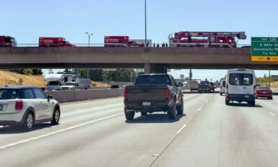 Fire trucks lined up on an overpass above a busy multi-lane highway as cars pass beneath them on the freeway.