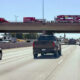 Fire trucks lined up on an overpass above a busy multi-lane highway as cars pass beneath them on the freeway.