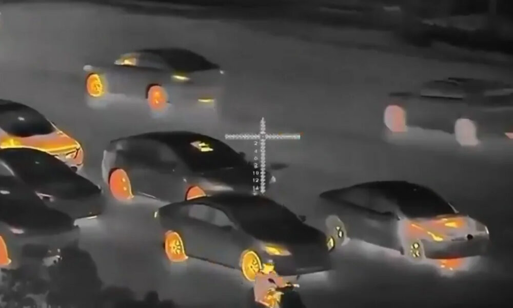 Night-vision style scene of multiple cars in a street chase, with orange-lit rims and a crosshair reticle overlaid in the center of the frame.