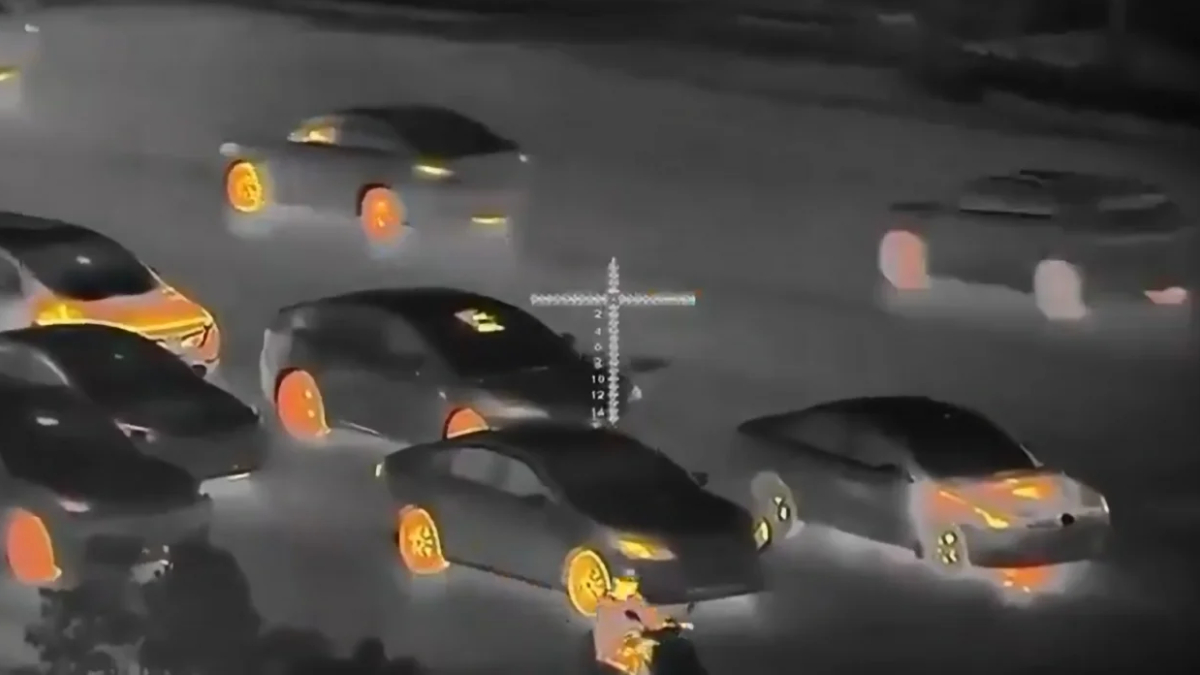 Night-vision style scene of multiple cars in a street chase, with orange-lit rims and a crosshair reticle overlaid in the center of the frame.