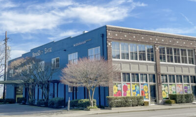 Blue brick storefront with large ground-floor windows showing colorful flower panels; bare trees line the sidewalk on a sunny day.