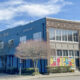 Blue brick storefront with large ground-floor windows showing colorful flower panels; bare trees line the sidewalk on a sunny day.