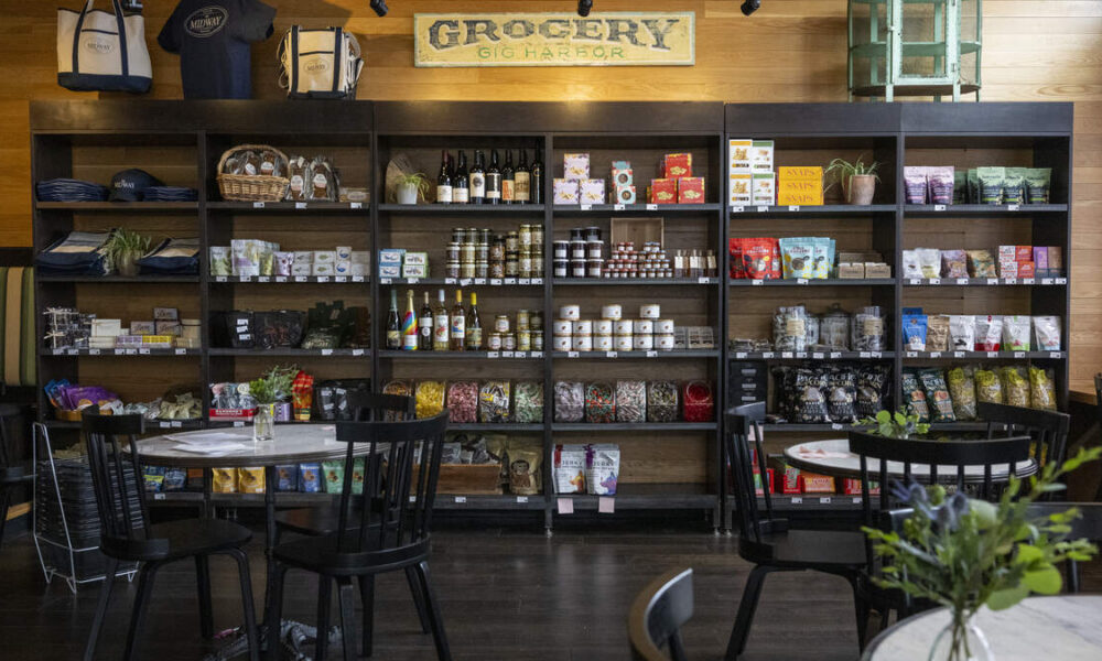 Shelves behind a dining area filled with jars, bottles, snacks and groceries in a cafe or market setting.