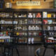 Shelves behind a dining area filled with jars, bottles, snacks and groceries in a cafe or market setting.