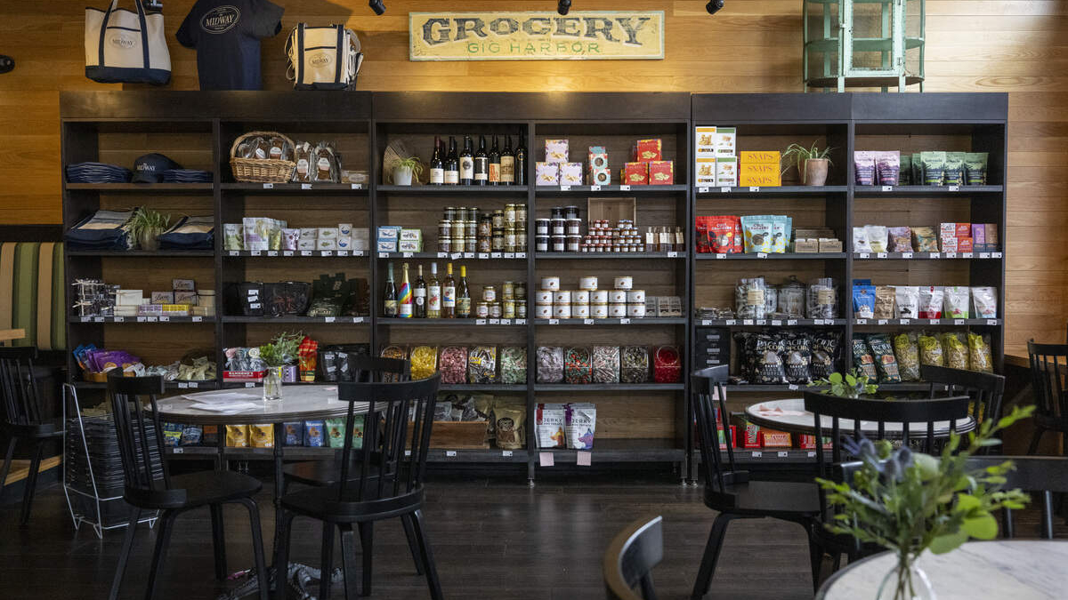 Shelves behind a dining area filled with jars, bottles, snacks and groceries in a cafe or market setting.