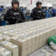 SWAT officers in tactical gear guard a currency processing area with huge stacks of bundled $100 bills and workers in the background.