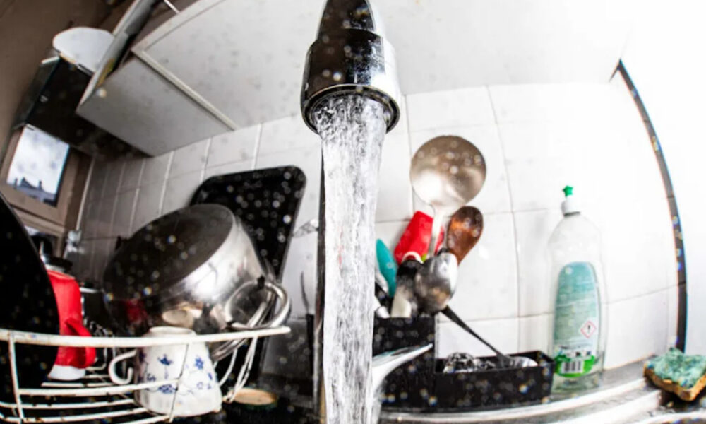 Water runs from a kitchen faucet into a sink full of dishes in a dish rack on the counter.