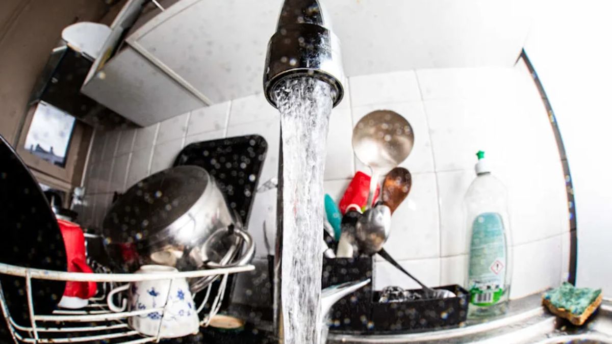 Water runs from a kitchen faucet into a sink full of dishes in a dish rack on the counter.