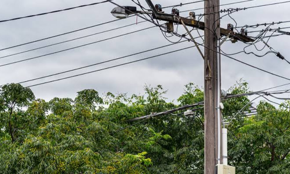 Utility pole with multiple power lines and a transformer, against a cloudy sky and green trees in the background.