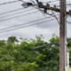 Utility pole with multiple power lines and a transformer, against a cloudy sky and green trees in the background.