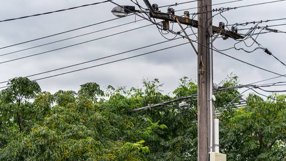 Utility pole with multiple power lines and a transformer, against a cloudy sky and green trees in the background.
