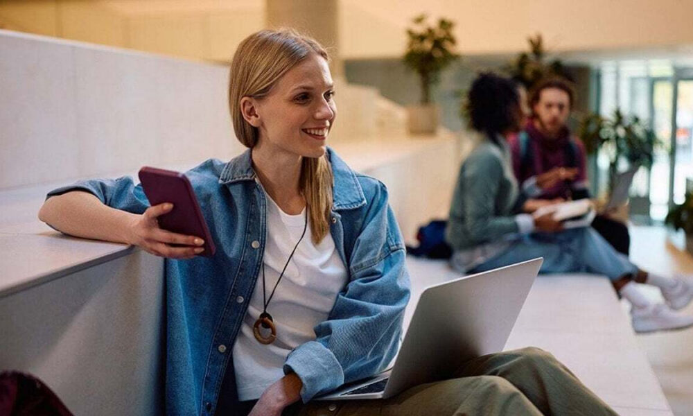 Young woman in a denim jacket sits on a stair ledge with a laptop and phone, smiling in a bright campus lobby.
