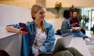Young woman in a denim jacket sits on a stair ledge with a laptop and phone, smiling in a bright campus lobby.