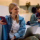 Young woman in a denim jacket sits on a stair ledge with a laptop and phone, smiling in a bright campus lobby.