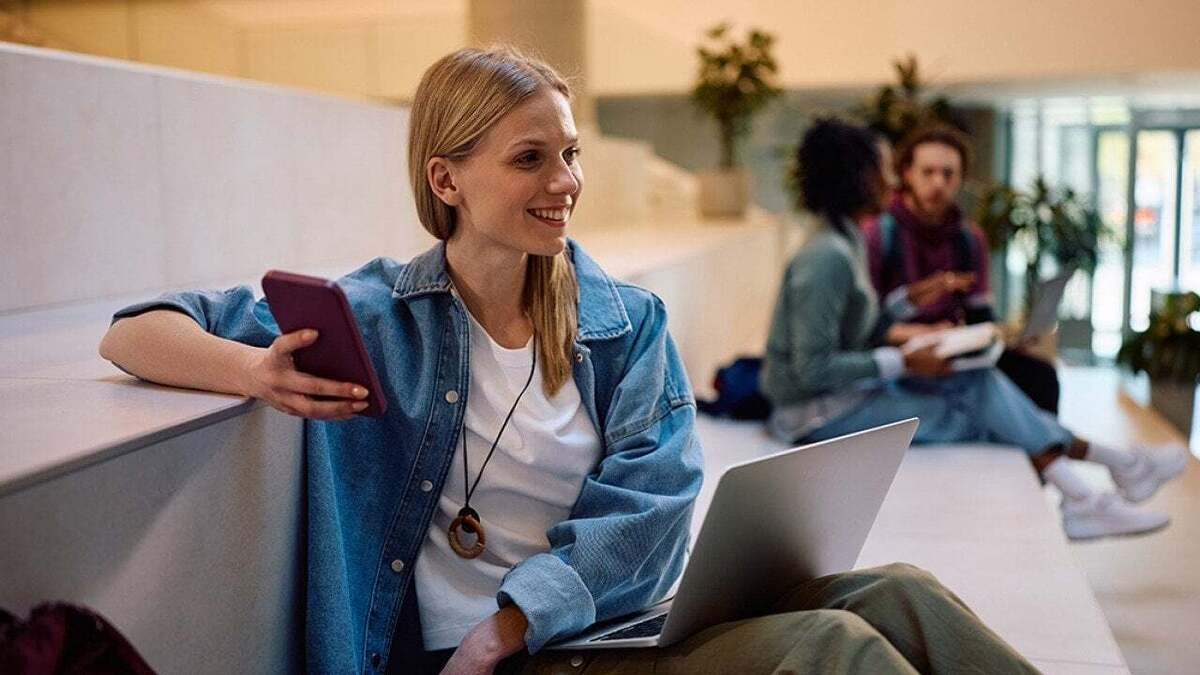 Young woman in a denim jacket sits on a stair ledge with a laptop and phone, smiling in a bright campus lobby.