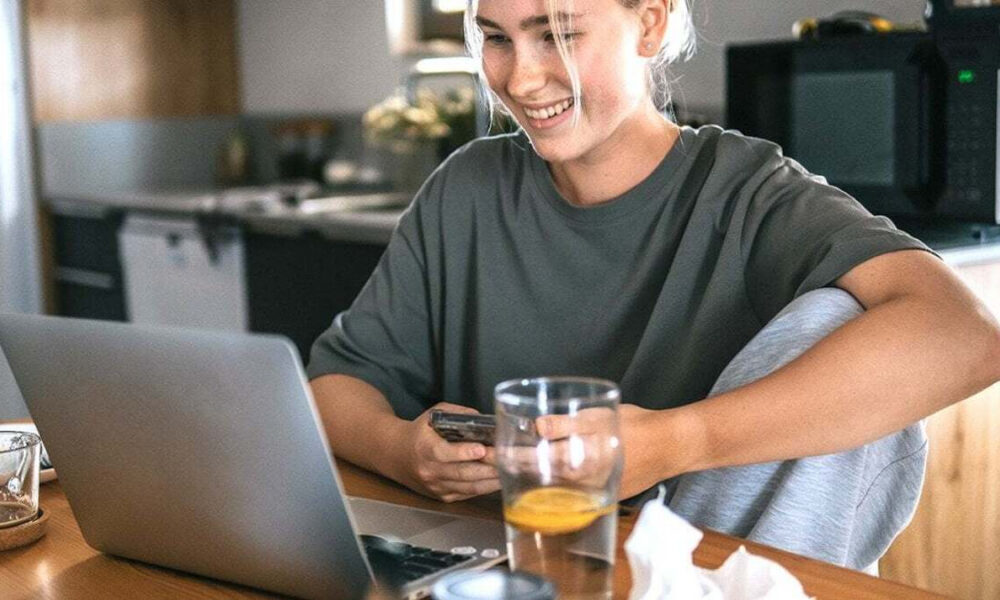Woman smiling at a laptop while seated at a kitchen table, holding a phone with a glass of water nearby.