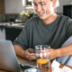 Woman smiling at a laptop while seated at a kitchen table, holding a phone with a glass of water nearby.