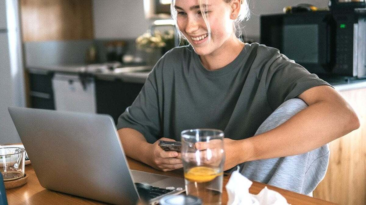 Woman smiling at a laptop while seated at a kitchen table, holding a phone with a glass of water nearby.