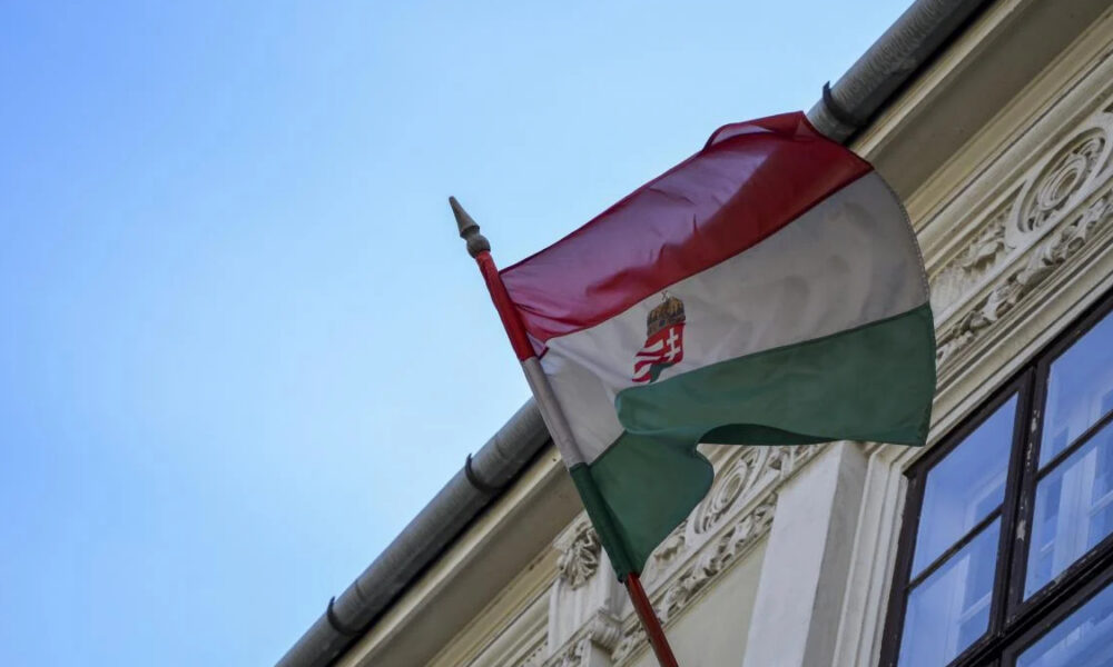 Hungarian flag waving on a flagpole beside an ornate building façade against a blue sky, showing red-white-green stripes and crest in the white stripe.