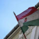 Hungarian flag waving on a flagpole beside an ornate building façade against a blue sky, showing red-white-green stripes and crest in the white stripe.
